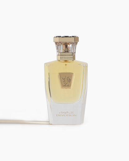A clear glass bottle of Devotion Parfum by Hind Al Oud features a gold cap, light yellow liquid, gold accents, and Arabic script. The oriental fragrance is showcased against a white background with its name "Devotion" in view.