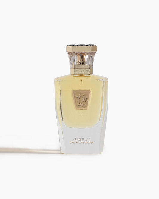 A clear glass bottle of Devotion Parfum by Hind Al Oud features a gold cap, light yellow liquid, gold accents, and Arabic script. The oriental fragrance is showcased against a white background with its name "Devotion" in view.