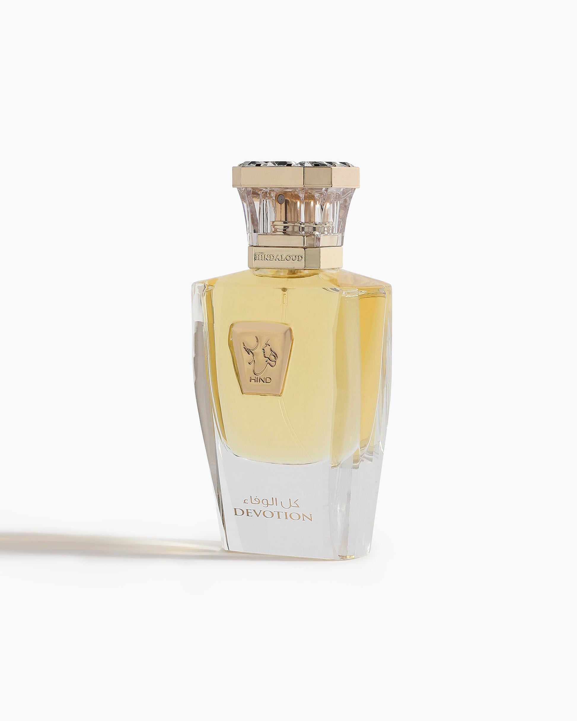 Devotion Parfum by Hind Al Oud is an elegant oriental perfume in a clear glass bottle with a gold cap, golden liquid, and an Arabic "DEVOTION" label. The white design features two hands forming a heart shape.