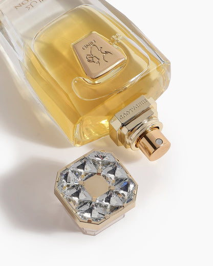 A close-up of Devotion Parfum by Hind Al Oud shows yellow liquid in a bottle with a gold label and a cap adorned with clear, faceted gems arranged in a circle, set against a white background.