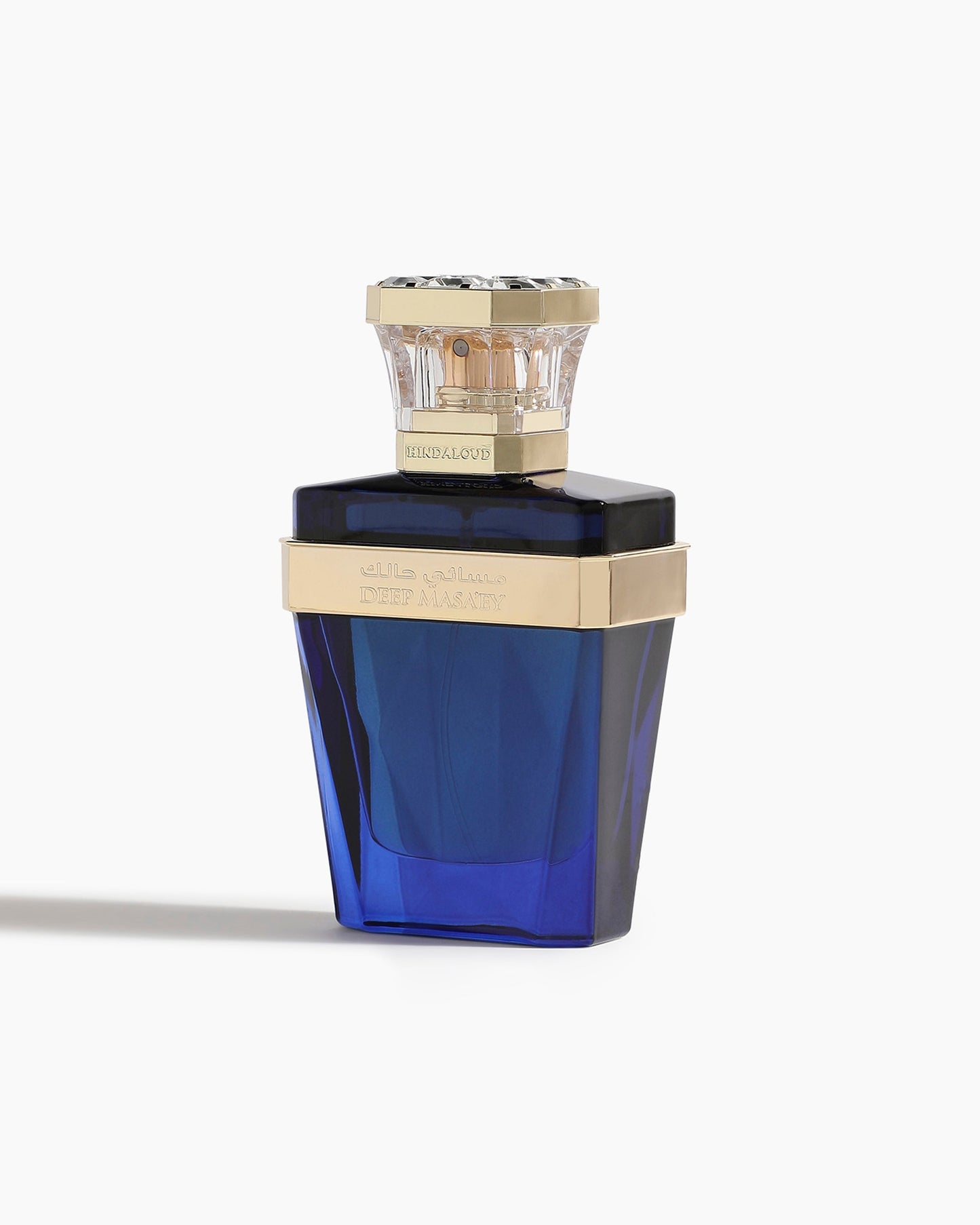 A deep blue, faceted bottle with a gold band and ornate cap, labeled Deep Masa'ey Parfum by Hind Al Oud, is set against a plain white background—an elegant display for its luxurious nutmeg and cashmere wood notes.