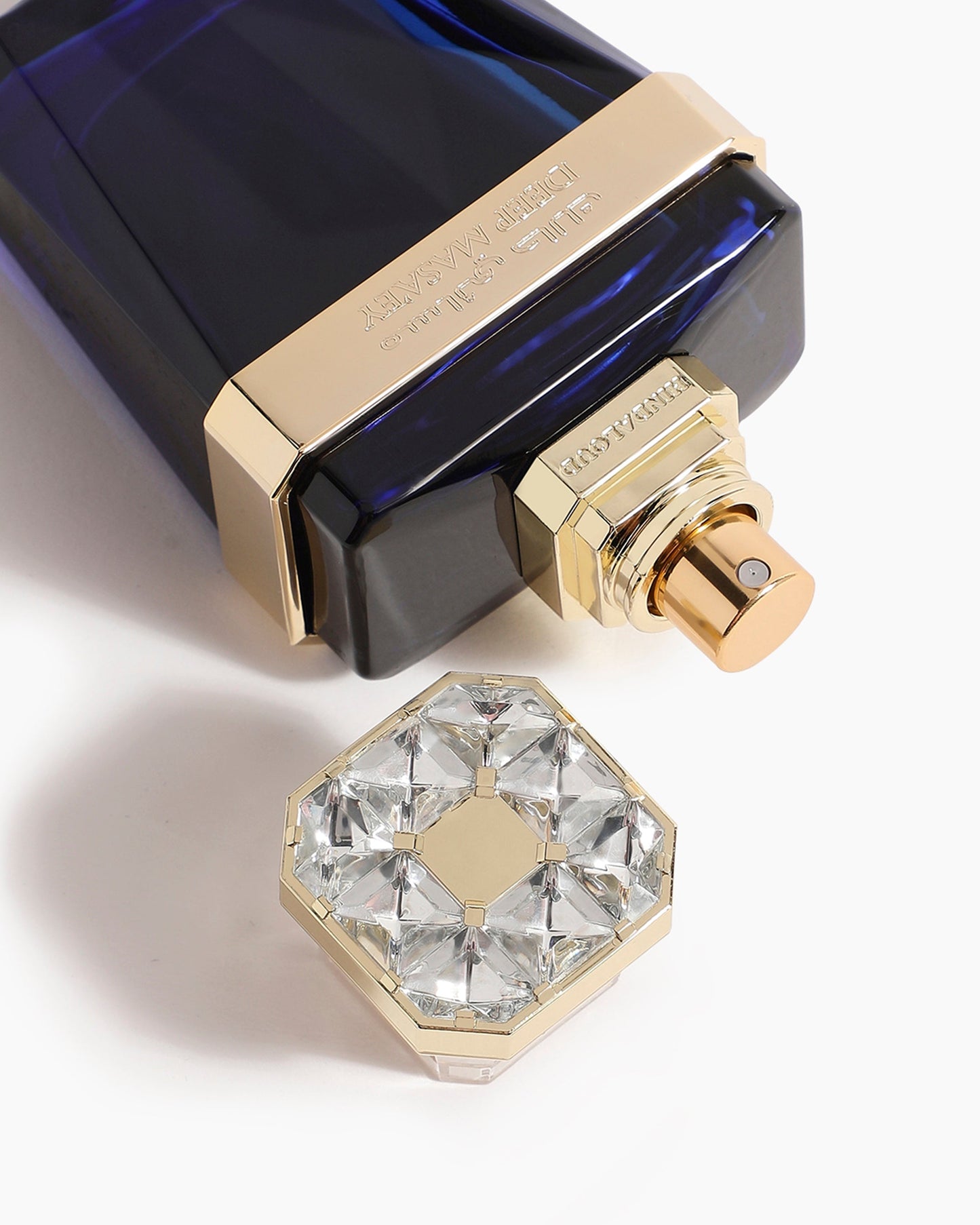 A close-up of the Deep Masa'ey Parfum by Hind Al Oud—a dark blue bottle with gold accents. Its cap, featuring clear crystals, rests beside it on a white surface, reflecting the elegance and richness within.