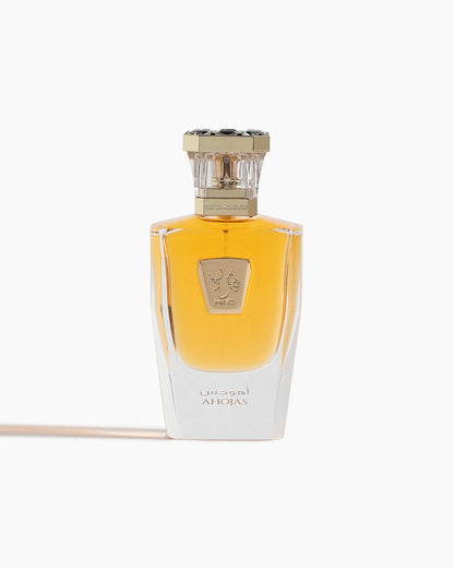 A transparent glass bottle with amber liquid, gold embossed emblem, and a gold-and-black cap. The label shows Ahojas in English and Arabic, identifying it as Ahojas Parfum by Hind Al Oud.
