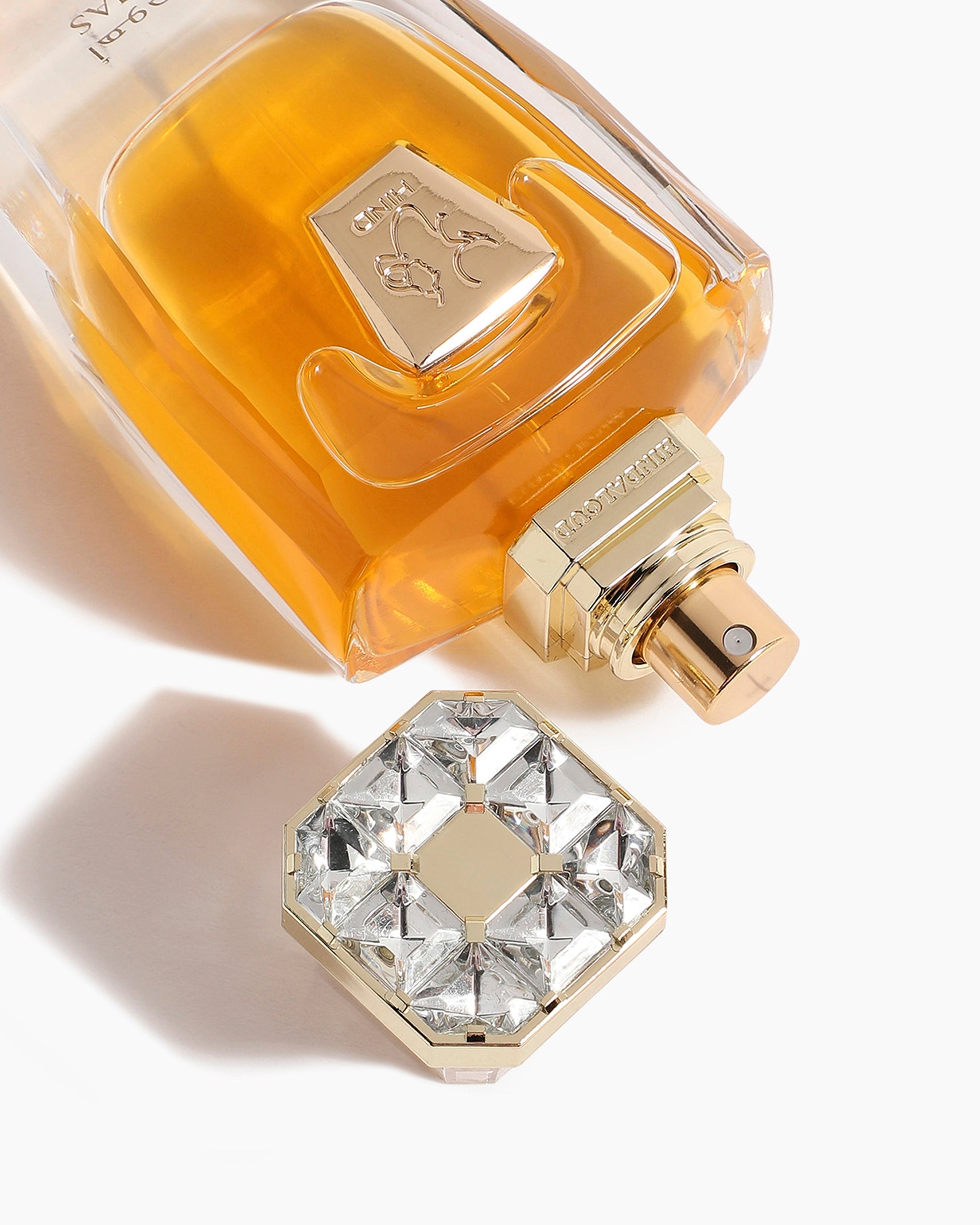 A gold-capped Ahojas Parfum - Hind Al Oud bottle from Hind Al Oud lies on its side, amber liquid visible. A gem-studded octagonal cap reflects light beside it, both elegantly suggesting the fig and tobacco scent within.