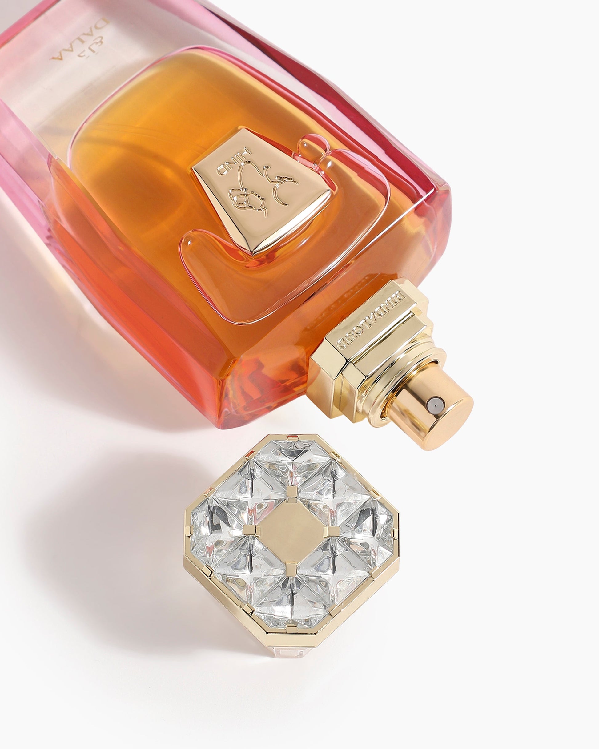 Dalaa Parfum by Hind Al Oud features a pink and amber gradient bottle with a gold spray nozzle, lying on its side. The gold cap with clear gem-like accents rests nearby on a white surface, suggesting the perfume's spicy floral scent.