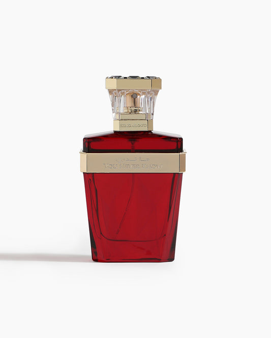 A red glass bottle of You Never Know Parfum by Hind Al Oud, with a gold and clear geometric cap and engraved text, sits against a white background—ideal for Amber Oud lovers.