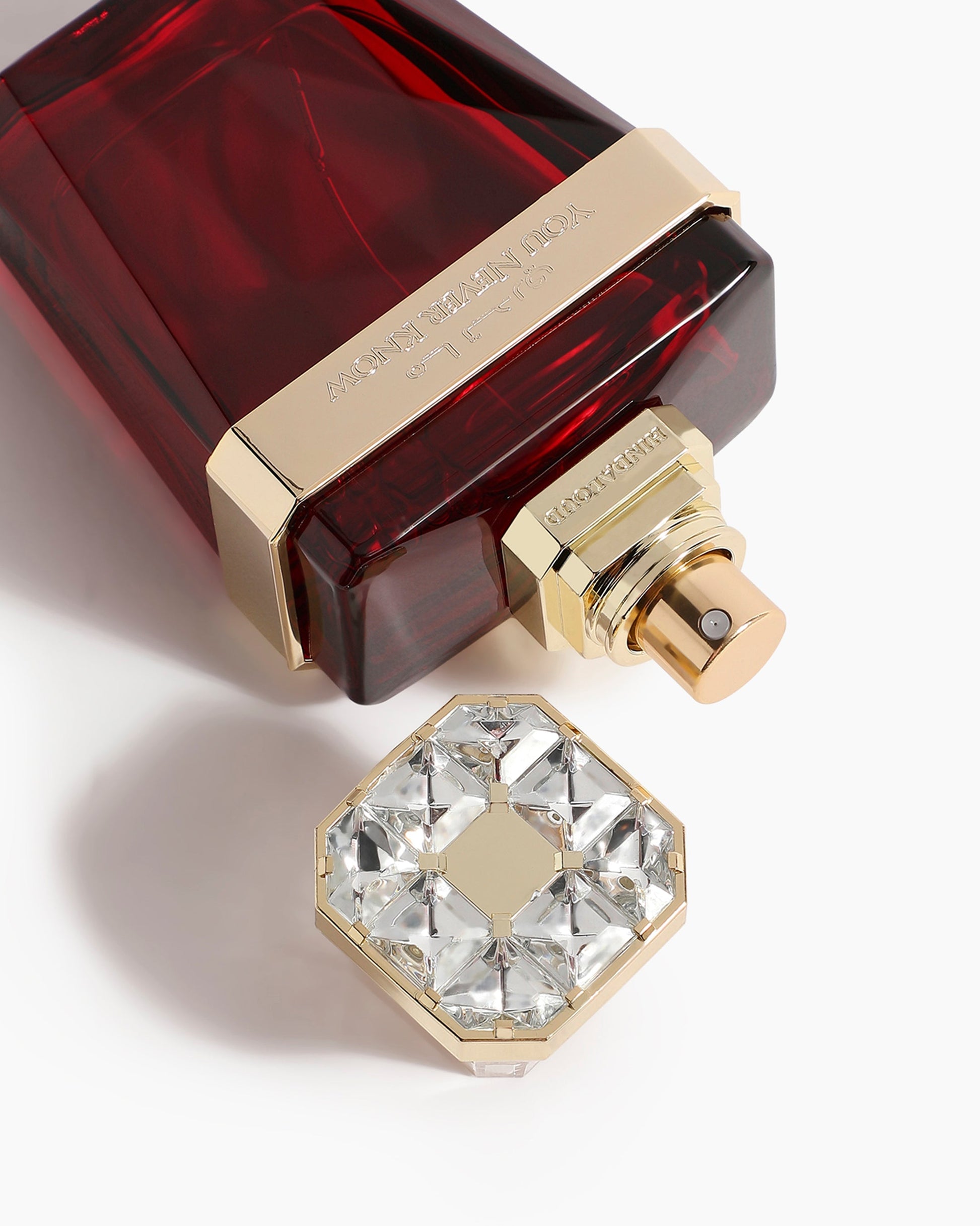 A close-up of the luxurious You Never Know Parfum by Hind Al Oud, featuring a dark red bottle with gold accents. The gem-studded cap rests beside it on a white background, while Amber Oud notes highlight its elegance.