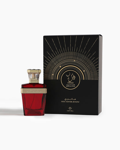 A red perfume bottle with a gold cap stands next to a black-and-gold box adorned with rays, Arabic script, and “You Never Know Parfum - Hind Al Oud.” Ideal for fans of luxurious Amber Oud scents by Hind Al Oud.