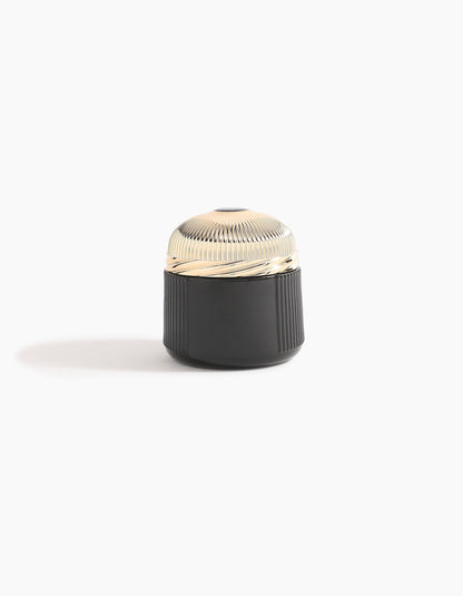 A small, round container with a ribbed, shiny gold lid and a black, textured base—echoing Agarwood luxury—Sheikh A Oud Mubakhar by Hind Al Oud rests on a plain white background with a soft shadow to the left.