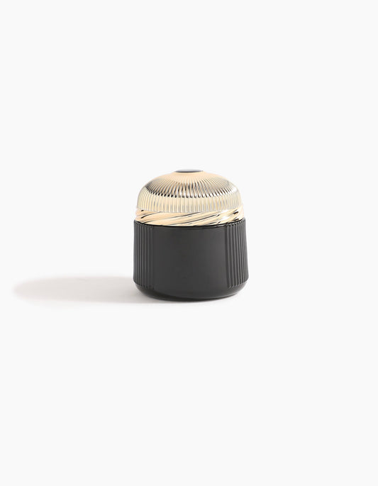 A small, round container with a ribbed, shiny gold lid and a black, textured base—echoing Agarwood luxury—Sheikh A Oud Mubakhar by Hind Al Oud rests on a plain white background with a soft shadow to the left.