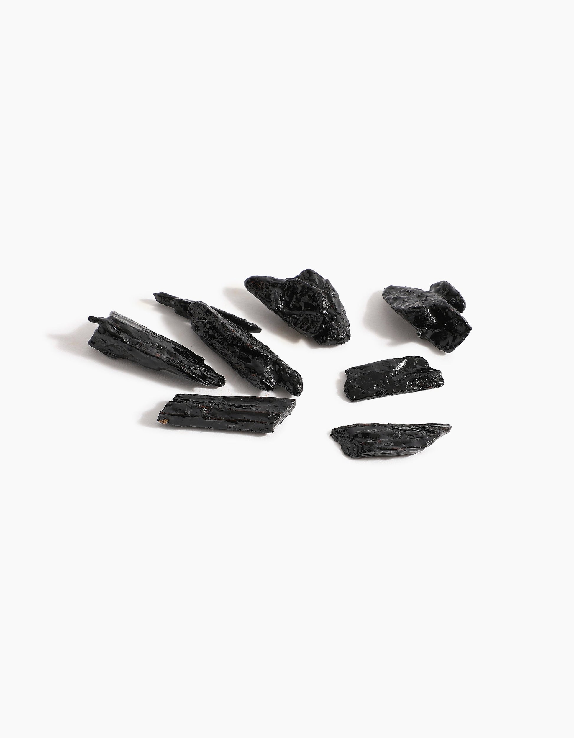 Eight pieces of rough, black tourmaline are scattered on a white surface, their jagged, shiny finish evoking the luxurious essence of Sheikh A Oud Mubakhar by Hind Al Oud.