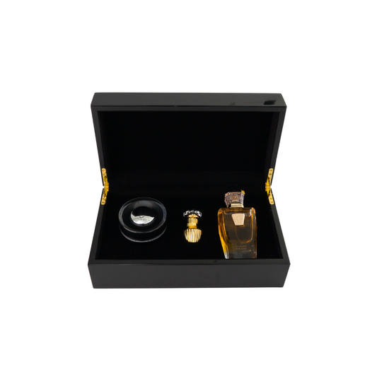 A black box with gold hinges, the Ahojas Collection Shield Edition Gift Box by Hind Al Oud, is open to reveal a small round container, an ornate bottle, and a larger rectangular bottle with amber liquid inside.