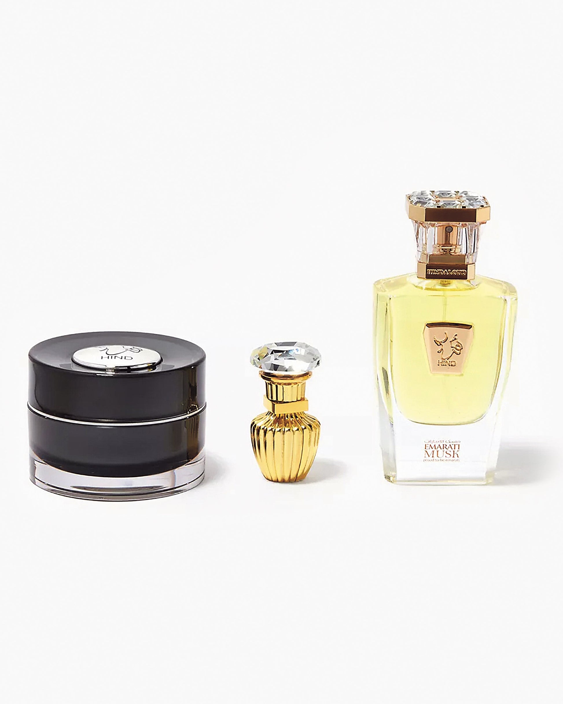 The Hind Al Oud Emarati Musk Trio Shield Edition Gift Box showcases three bottles: a round black jar with silver lid, a small gold decorative bottle in the center, and a tall rectangular glass bottle with a gold cap on the right.
