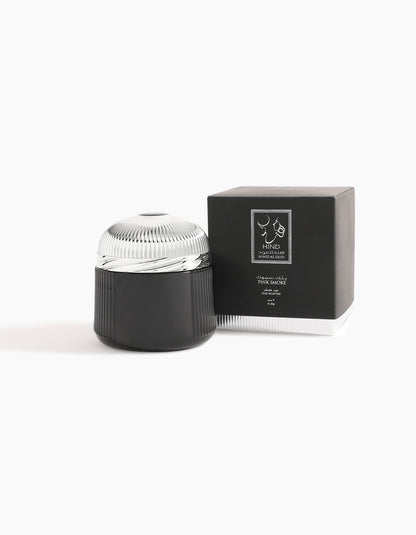 A black and silver cosmetic jar with a ridged lid sits next to a matching black box featuring a white label and the Hind Al Oud logo, evoking the refined notes of Pink Smoke Oud Muattar, set against a plain white background.