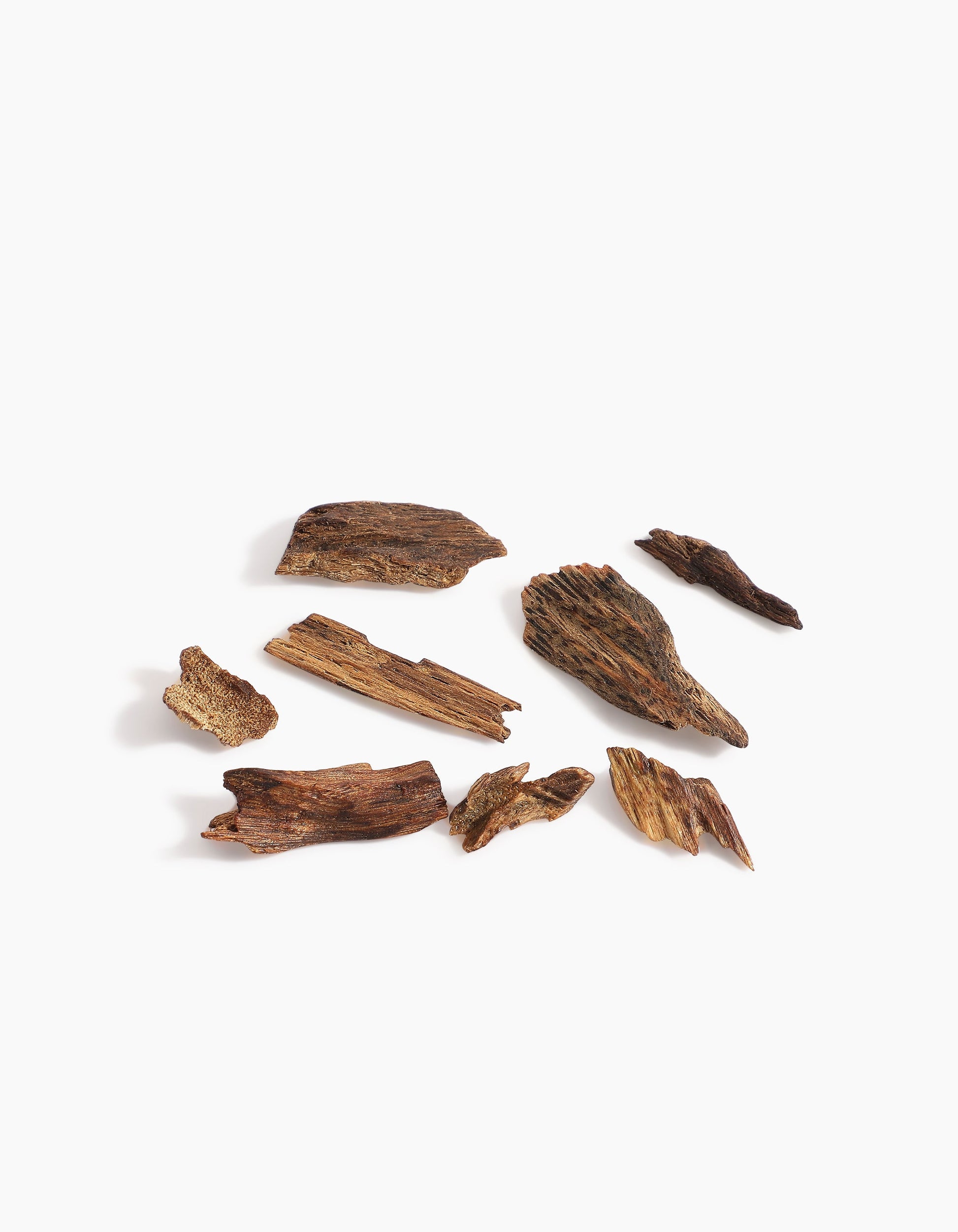 Several pieces of dark brown agarwood from Hind Al Oud’s Pink Smoke Oud Muattar are scattered on a white background, showcasing textured surfaces and natural grains with subtle notes similar to pink oud or gentle jasmine.