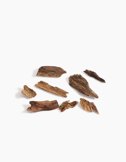 Several pieces of dark brown agarwood from Hind Al Oud’s Pink Smoke Oud Muattar are scattered on a white background, showcasing textured surfaces and natural grains with subtle notes similar to pink oud or gentle jasmine.