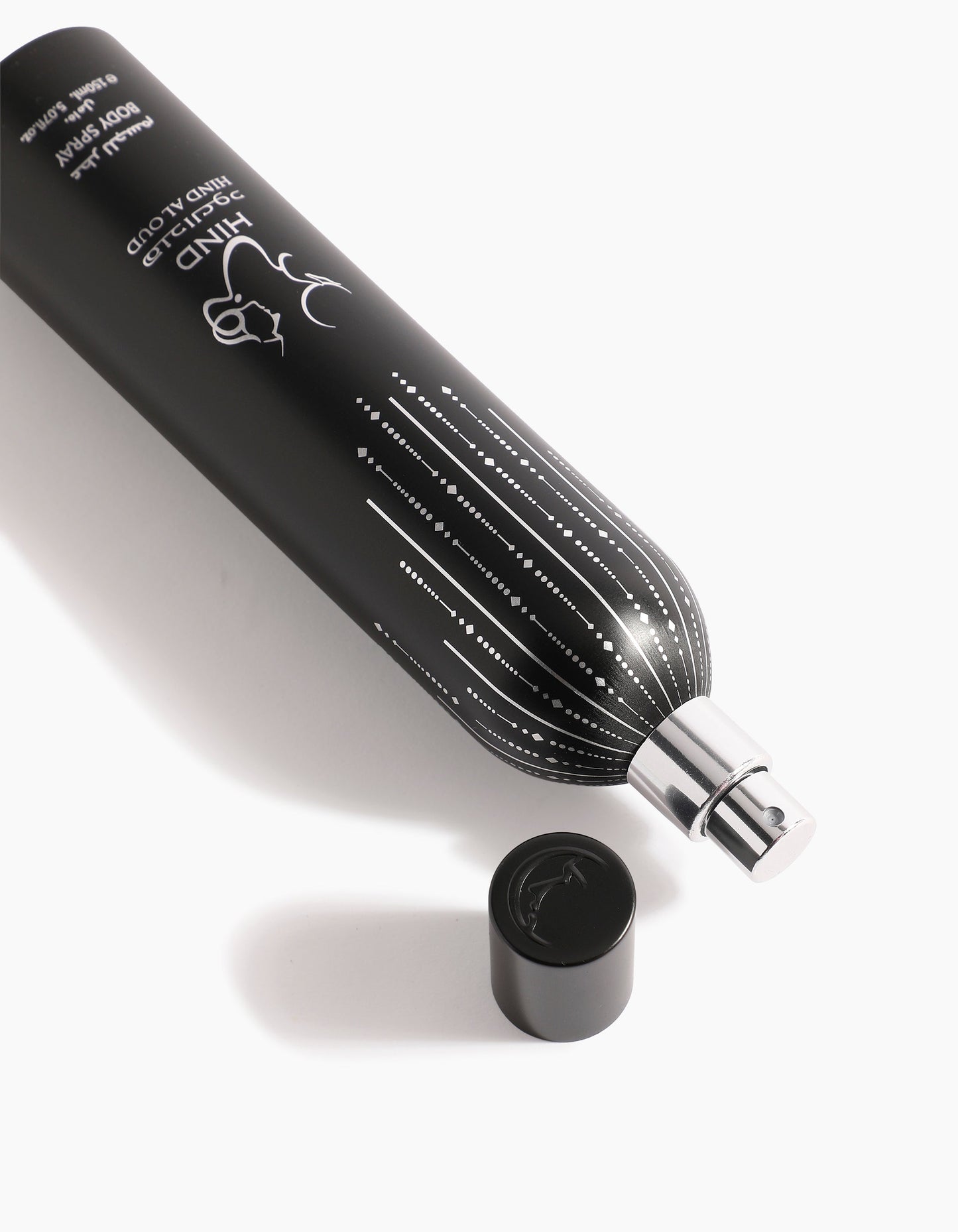 A black cylindrical spray bottle labeled Ahojas Body Spray by Hind Al Oud lies on its side with a silver nozzle and matching black cap nearby, showcasing an elegant design on a white background.