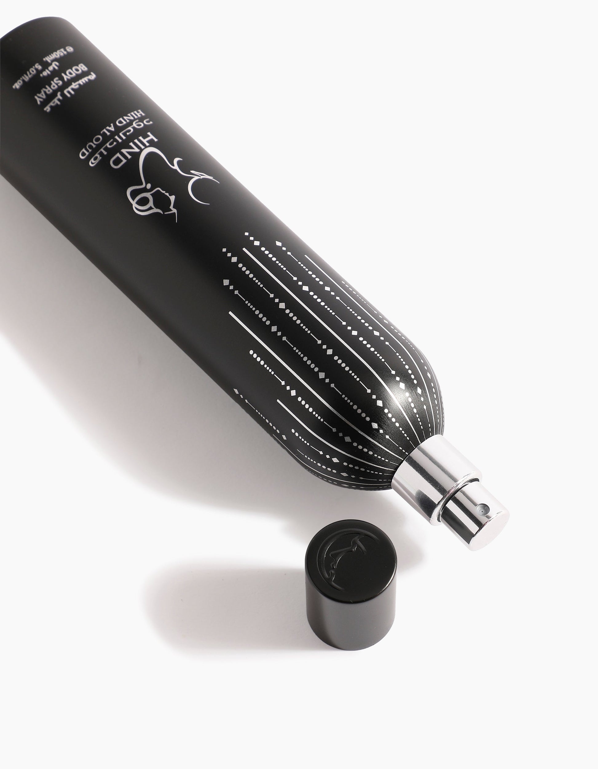 A black cylindrical spray bottle labeled Ahojas Body Spray by Hind Al Oud lies on its side with a silver nozzle and matching black cap nearby, showcasing an elegant design on a white background.