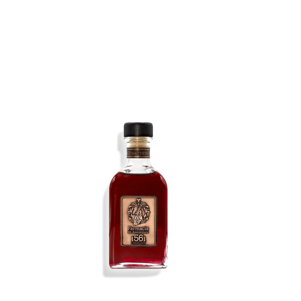 A small glass bottle with dark red liquid, black cap, and tan ornate label reading “Farmacia SS. Annunziata 1561 Vinattieri DIFFUSER,” resembles a diffuser with black sticks, suggesting a fruity wine scent. Displayed on a white background.