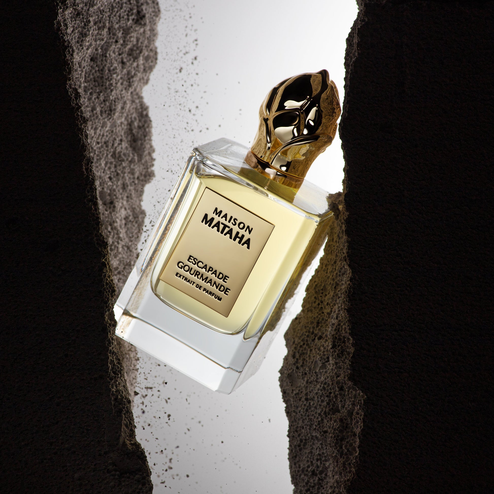 A bottle of Maison Mataha's Escapade Gourmande Extrait de Parfum, topped with a gold cap, hangs between rugged dark rocks, illuminated by a bright background light.