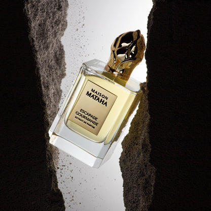 A bottle of Maison Mataha's Escapade Gourmande Extrait de Parfum, topped with a gold cap, hangs between rugged dark rocks, illuminated by a bright background light.