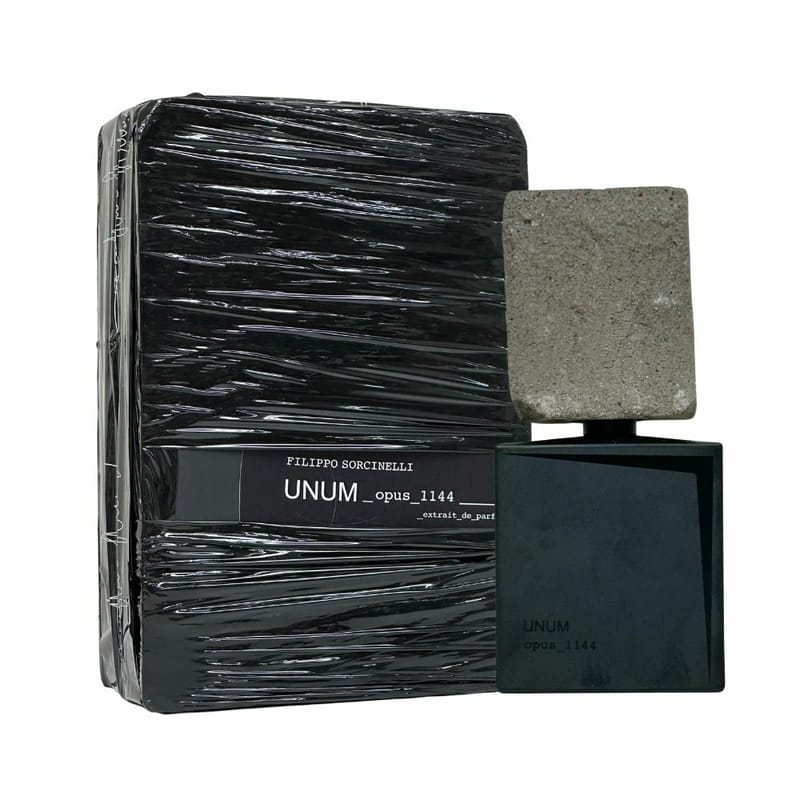 A bottle of UNUM OPUS 1144 "piece 1144" Eau de Parfum by Filippo Sorcinelli, featuring a rectangular stone-like cap, stands beside its black packaging, evoking sandalwood and white moss in an alluring fragrance experience.