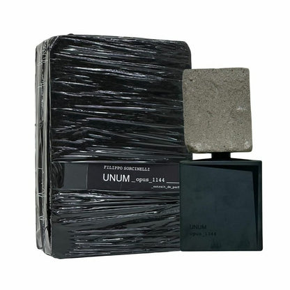 A bottle of UNUM OPUS 1144 "piece 1144" Eau de Parfum by Filippo Sorcinelli, featuring a rectangular stone-like cap, stands beside its black packaging, evoking sandalwood and white moss in an alluring fragrance experience.