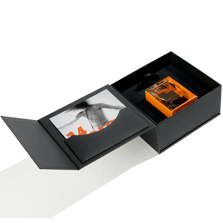 A black open gift box holds an orange 14 Hour Dream Eau de Parfum bottle by Jusbox, a patchouli scent by Antoine Lie, with a black-and-white photo of someone’s back on the inner lid.