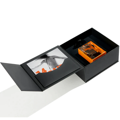 A black open gift box holds an orange 14 Hour Dream Eau de Parfum bottle by Jusbox, a patchouli scent by Antoine Lie, with a black-and-white photo of someone’s back on the inner lid.