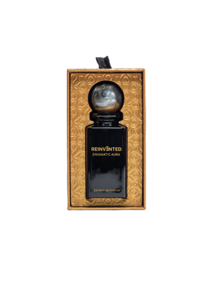 A black REINVĒNTED ENIGMATIC AURA Extrait de Parfum bottle with a round marbled cap sits in a textured gold box with circular patterns, showcasing vegan, cruelty-free elegance.