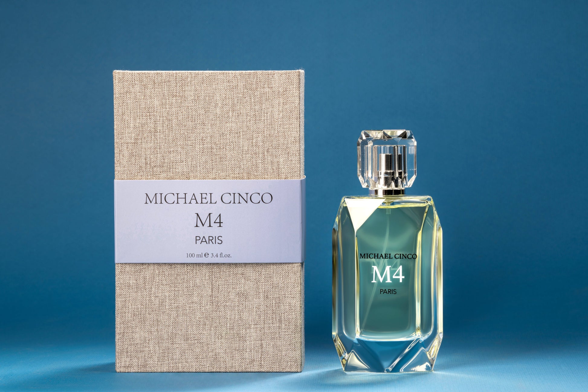 A faceted bottle of Fragrapedia Haus M4 Eau de Parfum by Michael Cinco, with marine notes, stands beside its textured beige box and blue label against a solid blue background, reflecting light elegantly.