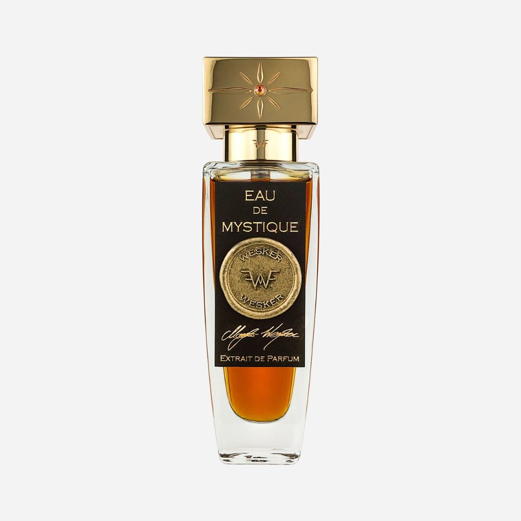 A clear glass Wesker Eau De Mystique Eau de Parfum bottle, filled with amber liquid, features a black label with a gold medallion, the Wesker logo, hints of Sicilian Blood Oranges, and a gold cap topped with a decorative star.