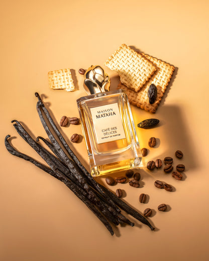The Café des Délices Extrait de Parfum by Maison Mataha sits amid vanilla beans, coffee beans, biscuits, and a single tonka bean on a beige background—evoking the richness of this gourmand fragrance.