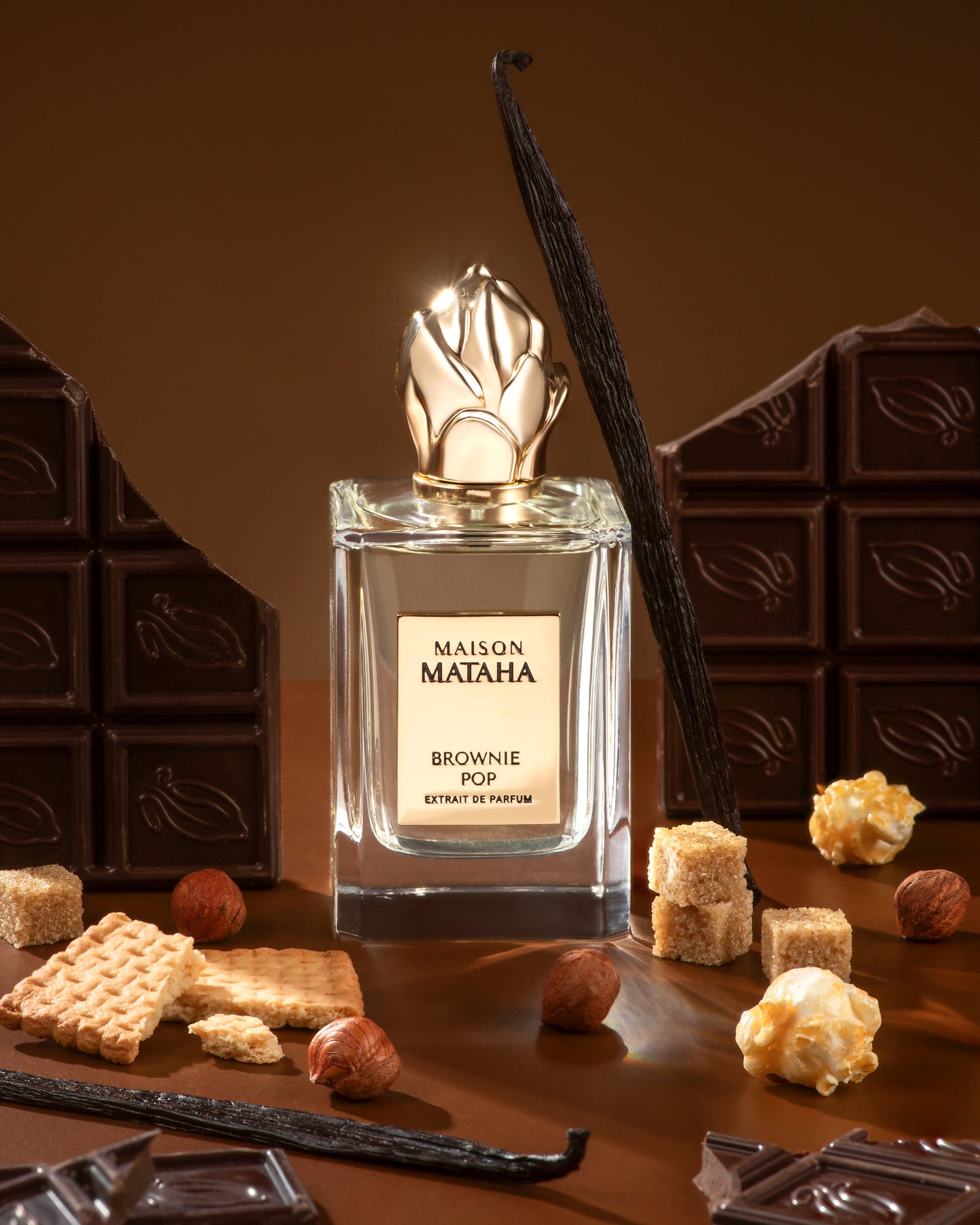 A bottle of Fragrapedia Haus Brownie Pop Extrait de Parfum by Maison Mataha, a decadent brownie scent, is displayed on a brown background with chocolate bars, vanilla beans, sugar cubes, hazelnuts, popcorn, and cookies.