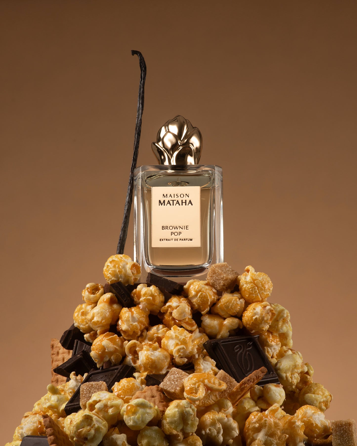 A bottle of Fragrapedia Haus Brownie Pop Extrait de Parfum by Maison Mataha rests on caramel popcorn, chocolate, and wafer cookies, with vanilla bean in the background—capturing a decadent brownie-inspired gourmand scent.