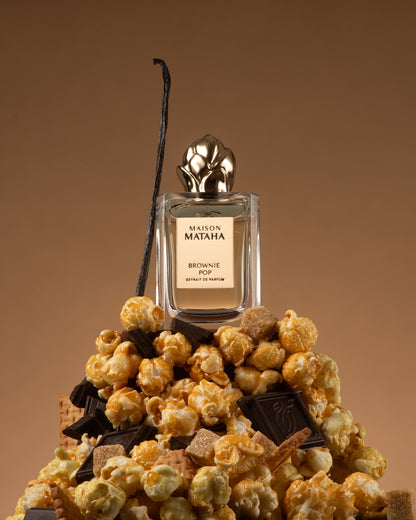 A bottle of Fragrapedia Haus Brownie Pop Extrait de Parfum by Maison Mataha rests on caramel popcorn, chocolate, and wafer cookies, with vanilla bean in the background—capturing a decadent brownie-inspired gourmand scent.