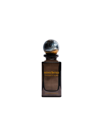 A dark brown glass bottle labeled ENIGMATIC AURA Extrait de Parfum - Reinvented by REINVĒNTED, with a round marble-like cap, is set against a white background—this vegan fragrance captures the mysterious essence of Enigmatic Aura.