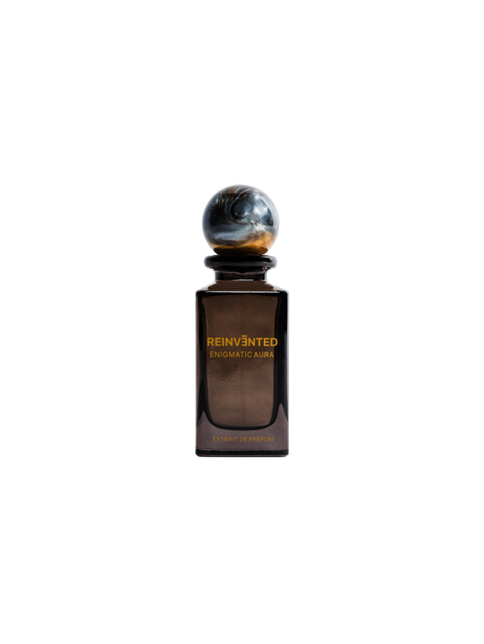 A dark brown glass bottle labeled ENIGMATIC AURA Extrait de Parfum - Reinvented by REINVĒNTED, with a round marble-like cap, is set against a white background—this vegan fragrance captures the mysterious essence of Enigmatic Aura.