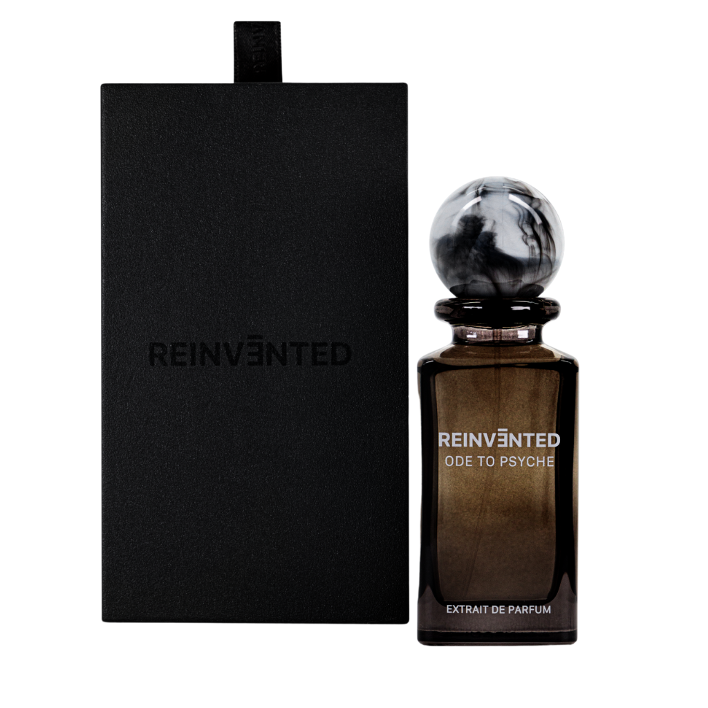 The limited edition ODE TO PSYCHE Extrait de Parfum - REINVENTED by REINVĒNTED features an elegant dark bottle with a round, marbled cap beside a matte black box embossed with the brand name, offering an alluring oriental woody scent.
