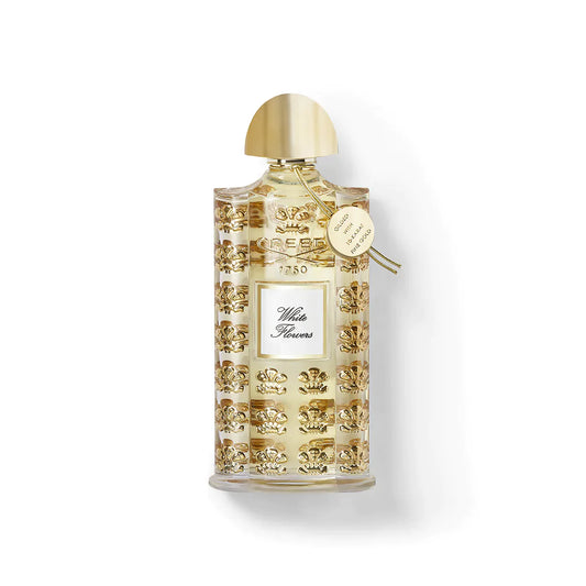 A luxurious clear glass bottle with gold accents, embossed designs, and a gold cap showcases CREED WHITE FLOWERS EDP by CREED. The elegant label and gold tag on the neck highlight this exclusive luxury fragrance.