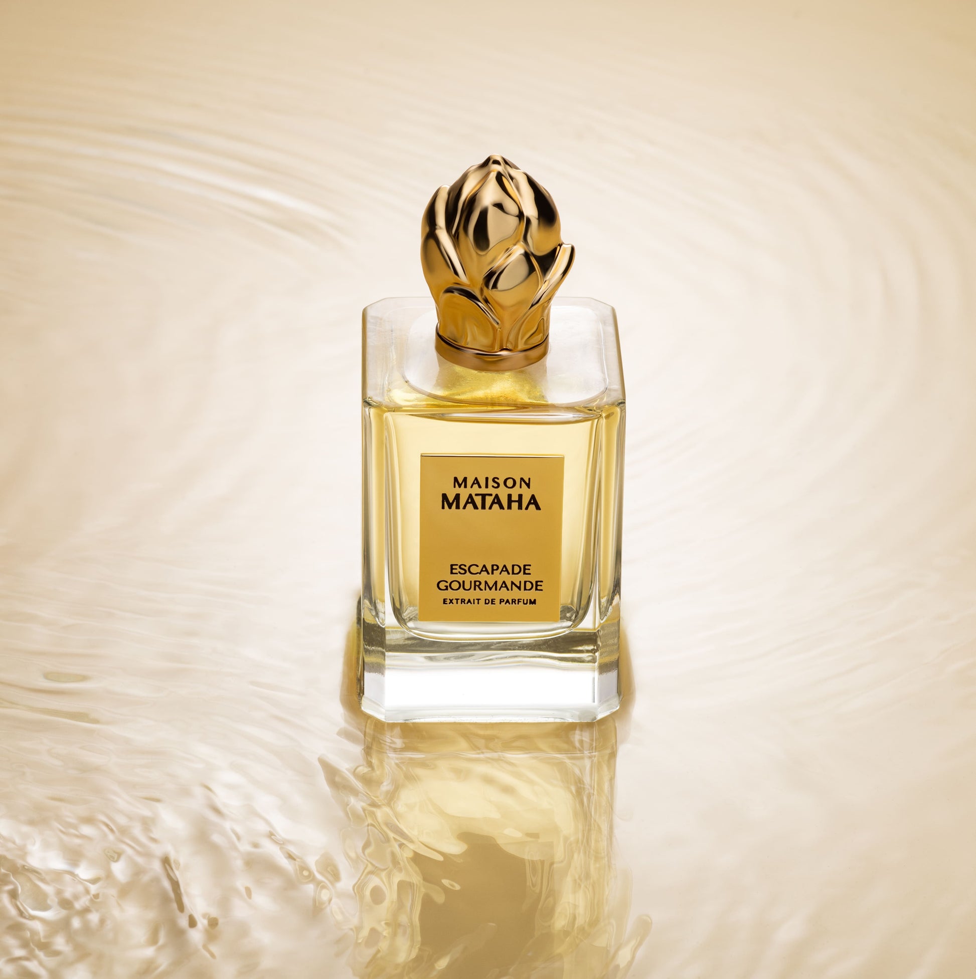 The Escapade Gourmande Extrait de Parfum by Maison Mataha, in a glass bottle with a gold cap on a golden rippled surface, captures the essence of vanilla and creamy crème brûlée.