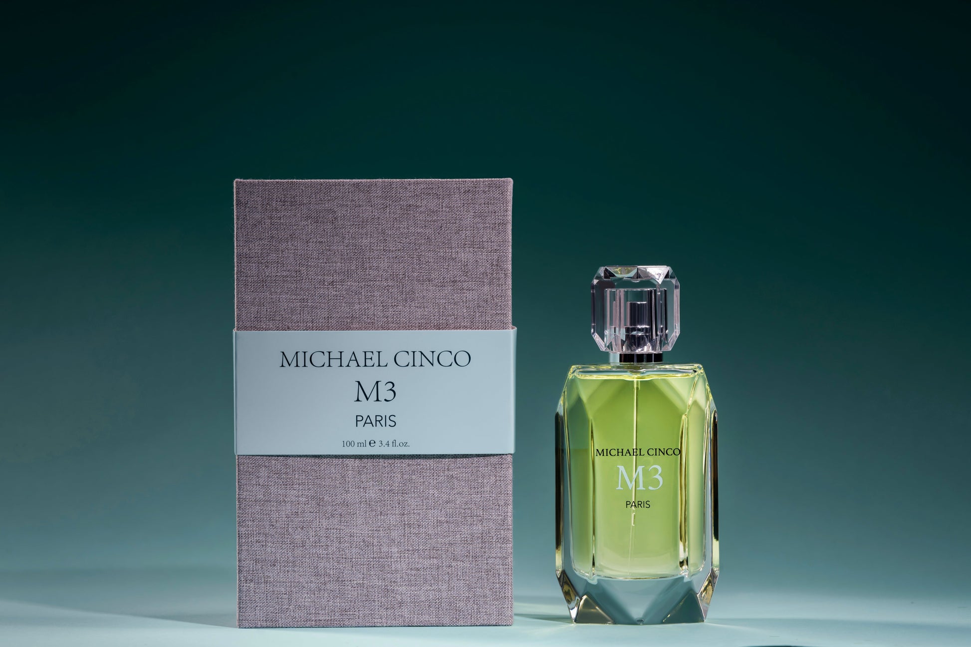 A bottle of Fragrapedia Haus M3 Eau de Parfum by Michael Cinco, with green liquid, stands beside its textured gray box on a dark teal background—an elegant women’s floral scent made with consciously sourced ingredients.