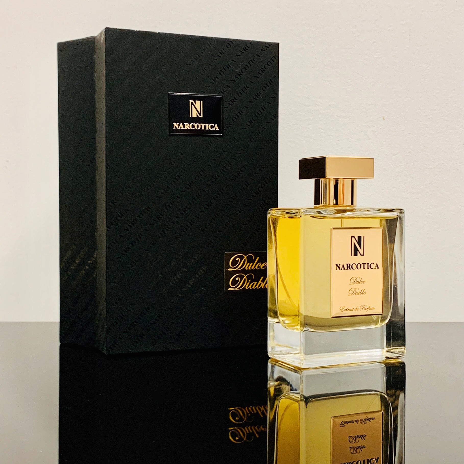 A bottle of Dulce Diablo Eau de Parfum by Narcotica with a gold cap stands beside its black and gold box on a reflective surface, set against a plain light background.