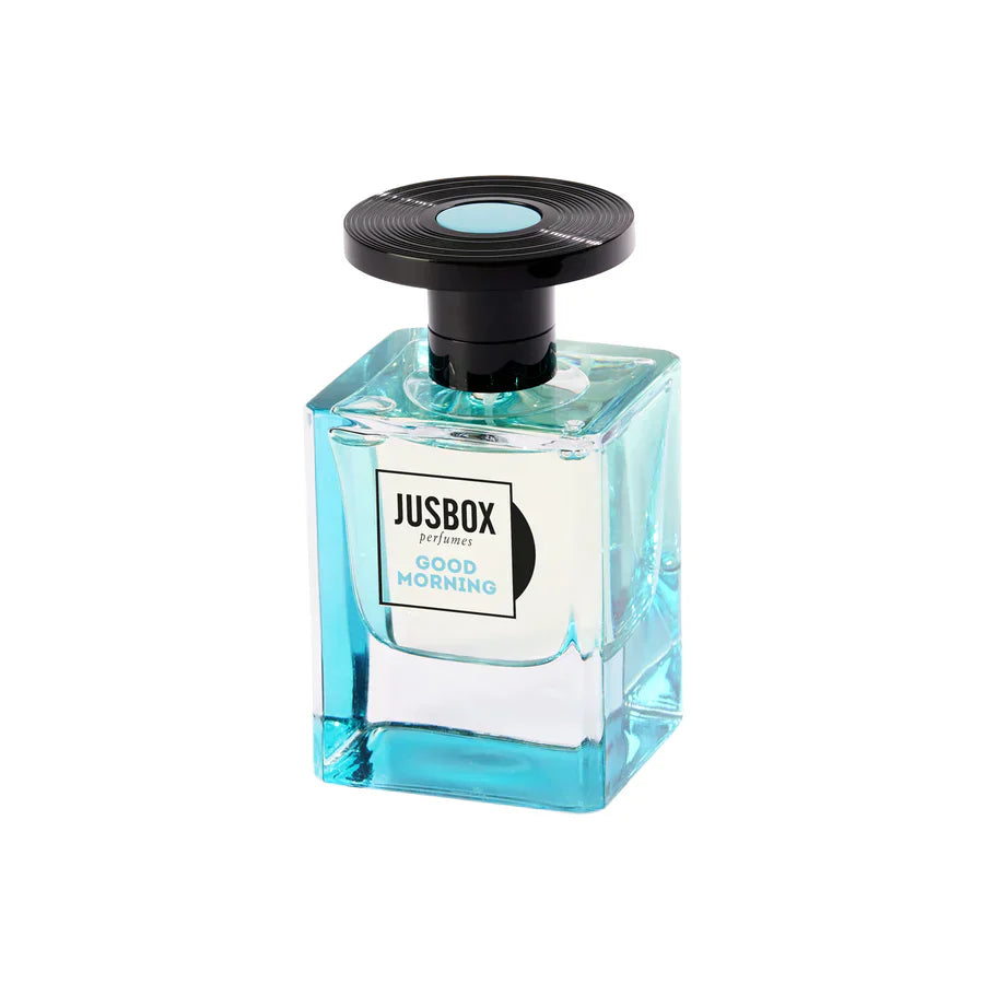 A clear rectangular bottle with light blue liquid, labeled Good Morning Eau de Parfum - JUSBOX by Jusbox, topped with a round black cap and blue accent—a refreshing fragrance with hints of magnolia.