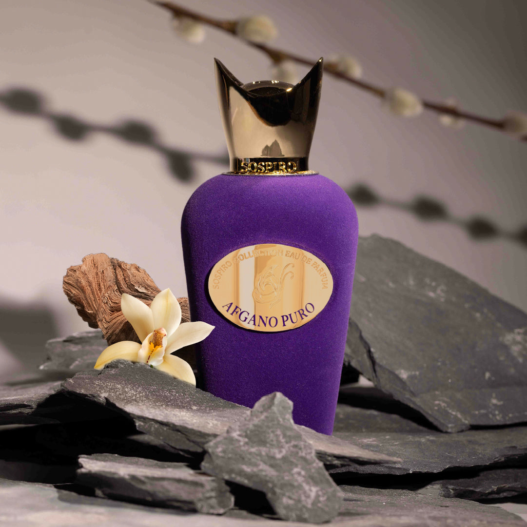 A purple, gold-capped Afgano Puro Eau de Parfum by SOSPIRO stands on gray stone with a white flower and wood, evoking the rich allure of cedar patchouli, against a blurred branch background.