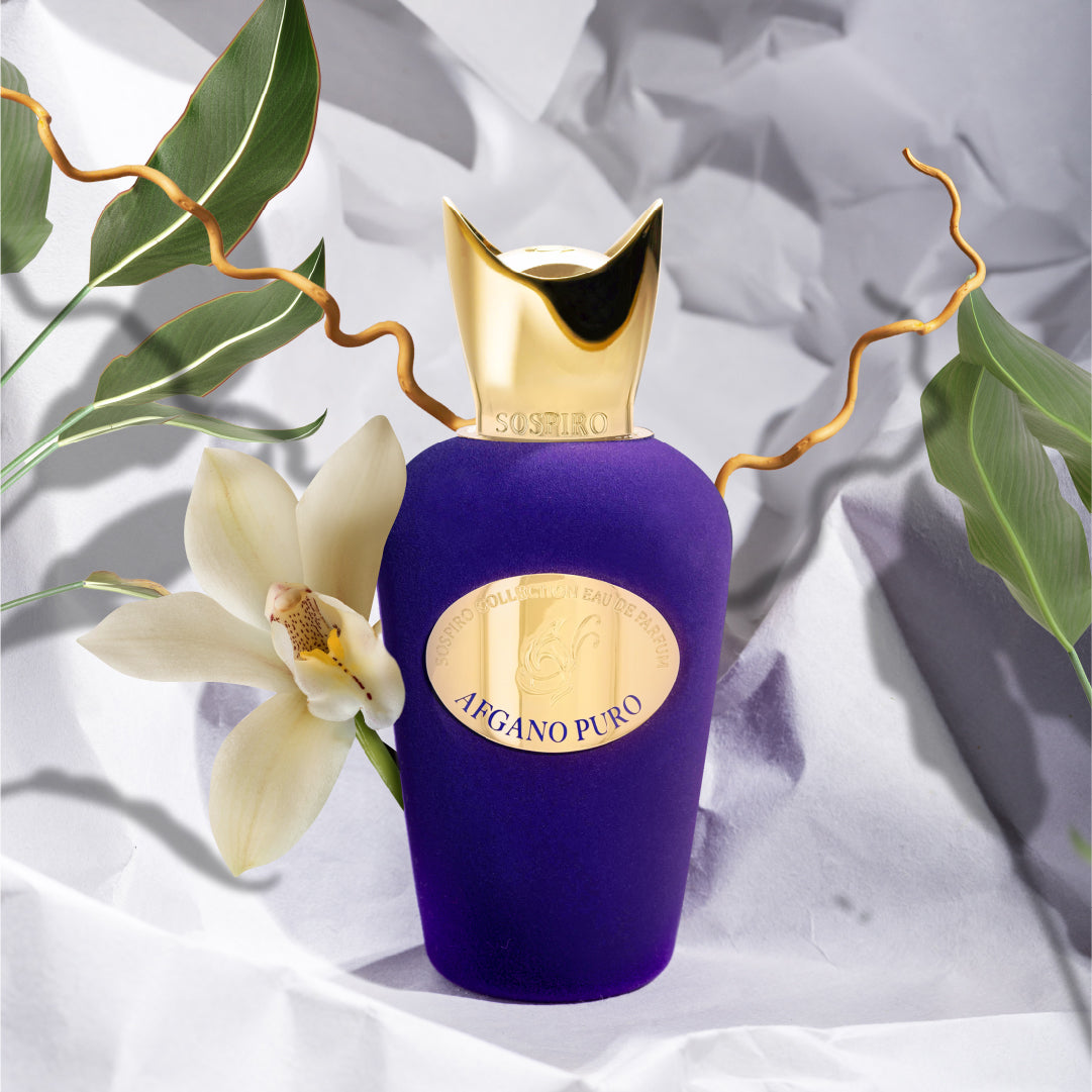 A purple and gold bottle of Afgano Puro Eau de Parfum by SOSPIRO is displayed with a white orchid and green leaves on a crumpled white background, featuring an elegant curved gold cap that suggests refined woody and jazz club fragrance notes.