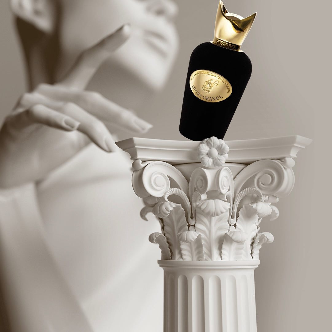 A black and gold Opera Grande Eau de Parfum by SOSPIRO rests on an ornate white Corinthian column, with a blurred statue in the background, evoking notes of Turkish Rose.