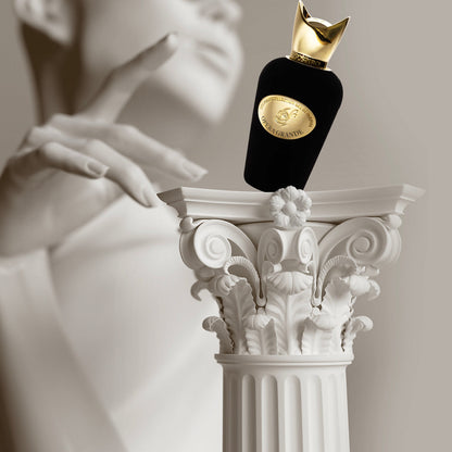 A black and gold Opera Grande Eau de Parfum by SOSPIRO rests on an ornate white Corinthian column, with a blurred statue in the background, evoking notes of Turkish Rose.