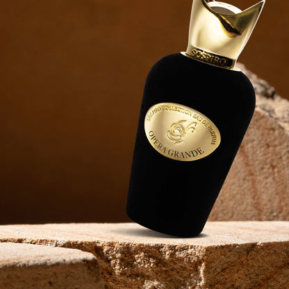 A black velvet bottle of Opera Grande Eau de Parfum by SOSPIRO, with a gold cap and oval label, stands on rough beige stone, evoking Turkish Rose notes against a blurred brown backdrop.