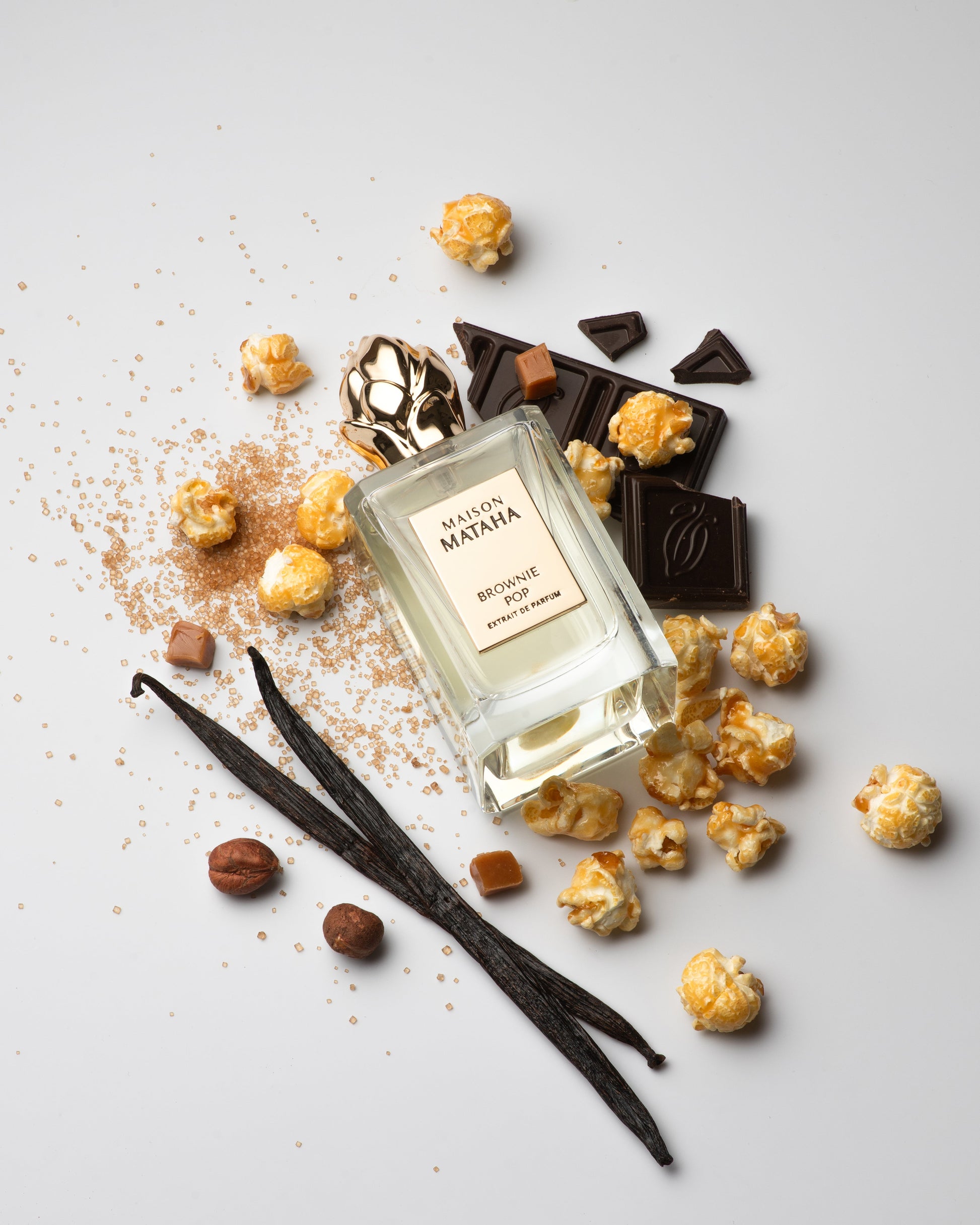 A bottle of Brownie Pop Extrait de Parfum by Fragrapedia Haus rests among popcorn, caramel, chocolate, vanilla beans, hazelnut, and brown sugar—capturing the deliciously decadent essence of a brownie-inspired gourmet scent.